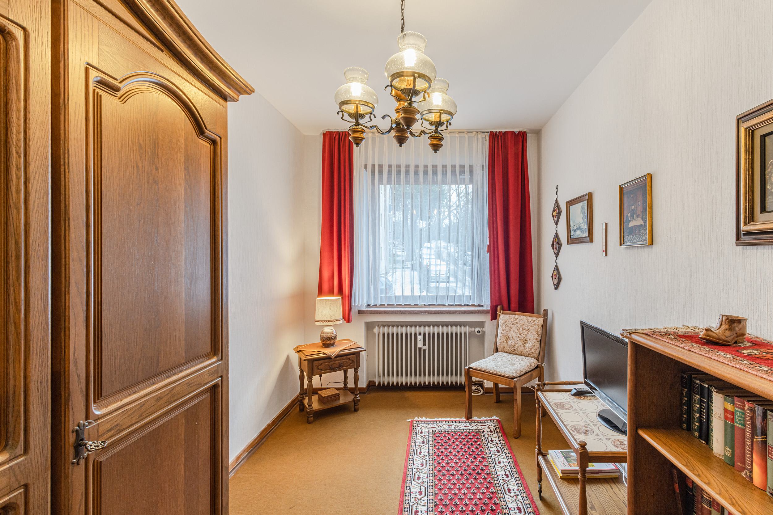 Kleines Wohnzimmer mit Holzschrank, Teppich, TV, Sessel und Fenster mit roten Vorhängen.