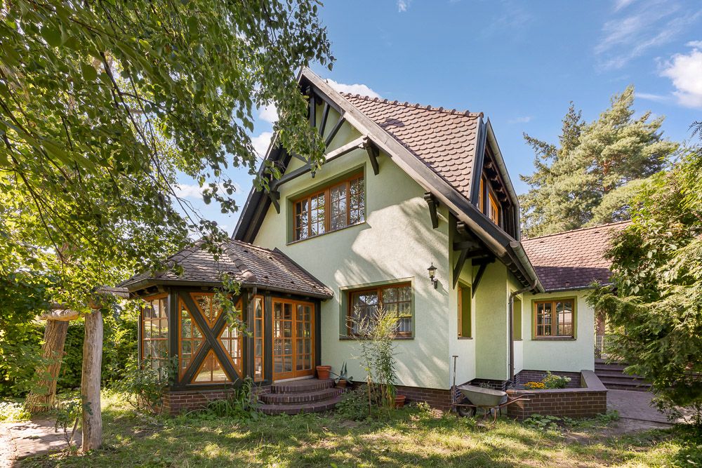 Zweistöckiges Haus mit Satteldach, Holzfenstern und Wintergarten, umgeben von Bäumen und Garten.
