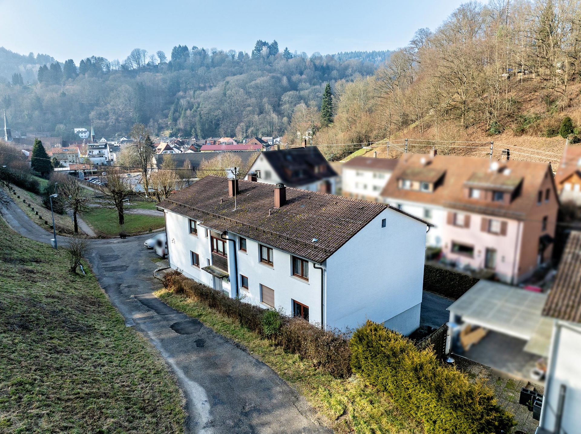 Zweistöckiges Wohnhaus mit braunem Dach an einer kleinen Straße, umgeben von Bäumen und anderen Häusern.