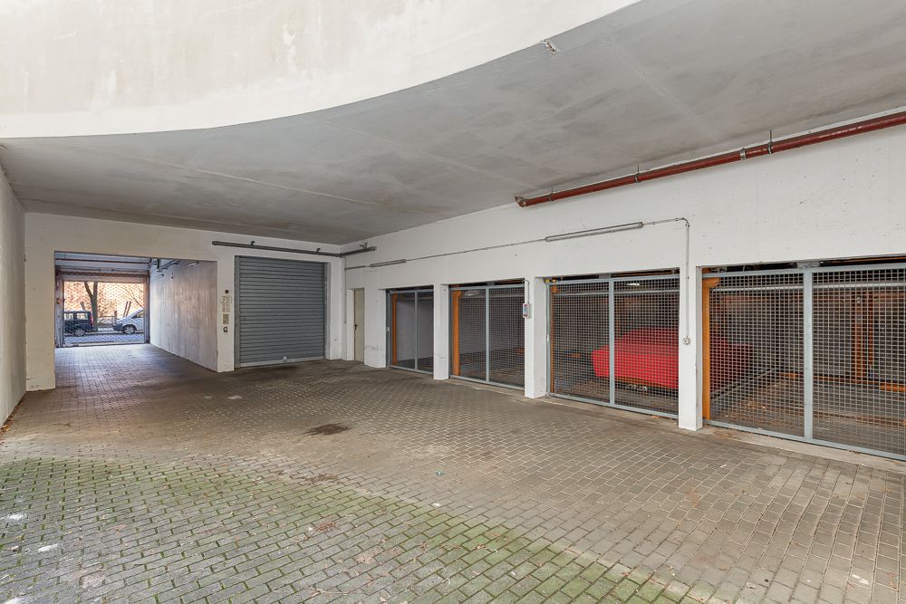 Überdachte Tiefgarageneinfahrt mit Gittertüren und parkenden Autos dahinter.