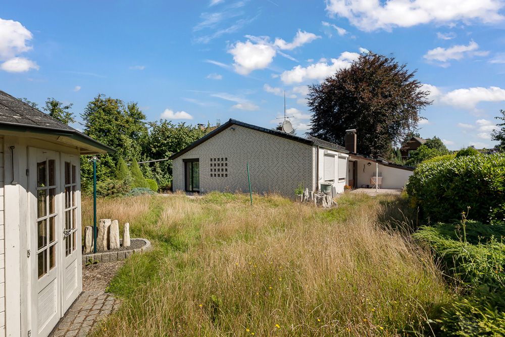 Garten mit hohem Gras, weißem Haus und Nebengebäude bei blauem Himmel.