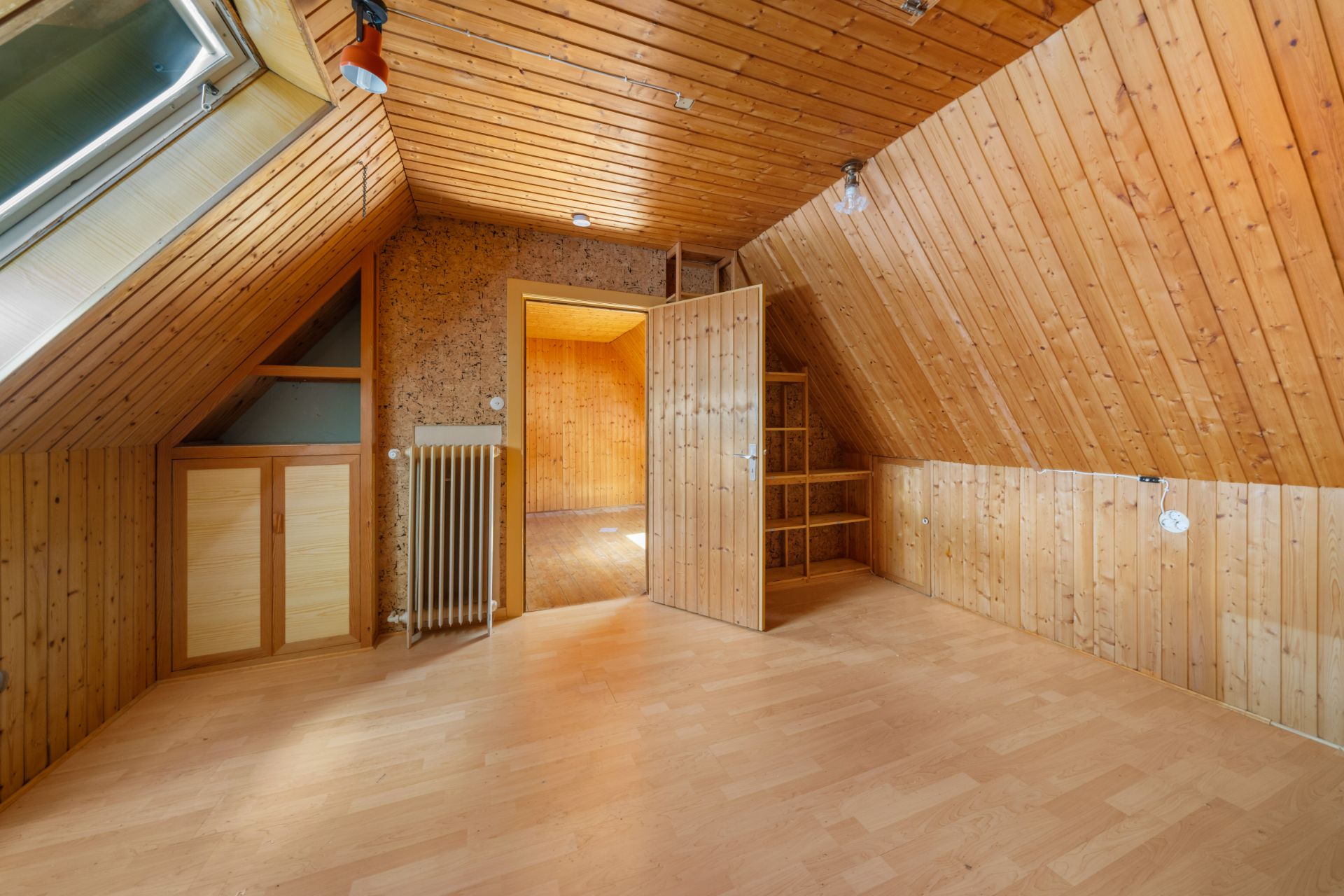 Dachzimmer mit Holzvertäfelung, Dachfenster, offenem Regal und Heizung.