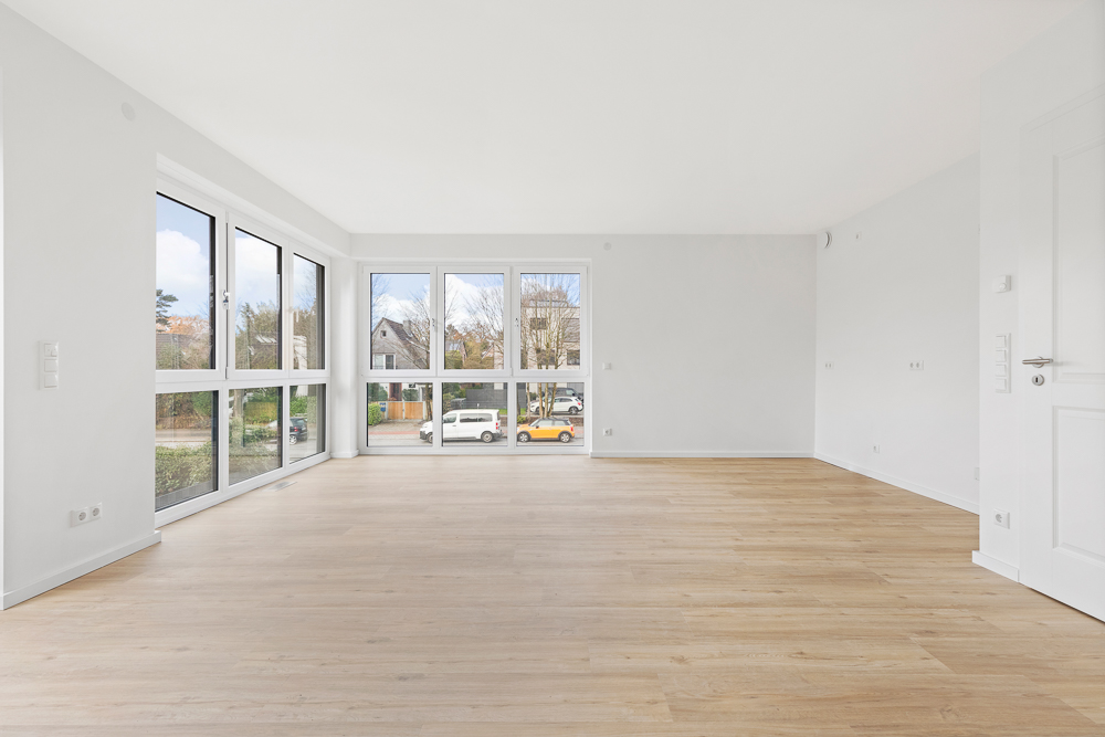 Helles, leeres Wohnzimmer mit großen Fenstern und Holzfußboden.