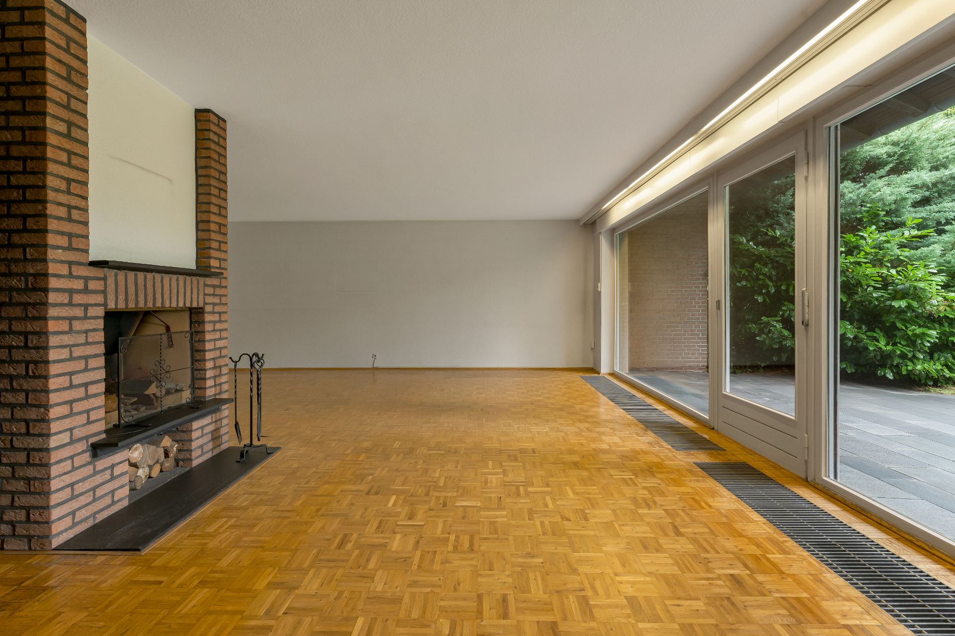 Großes, leeres Wohnzimmer mit Kamin, Parkettboden und bodentiefen Fenstern zum Garten.