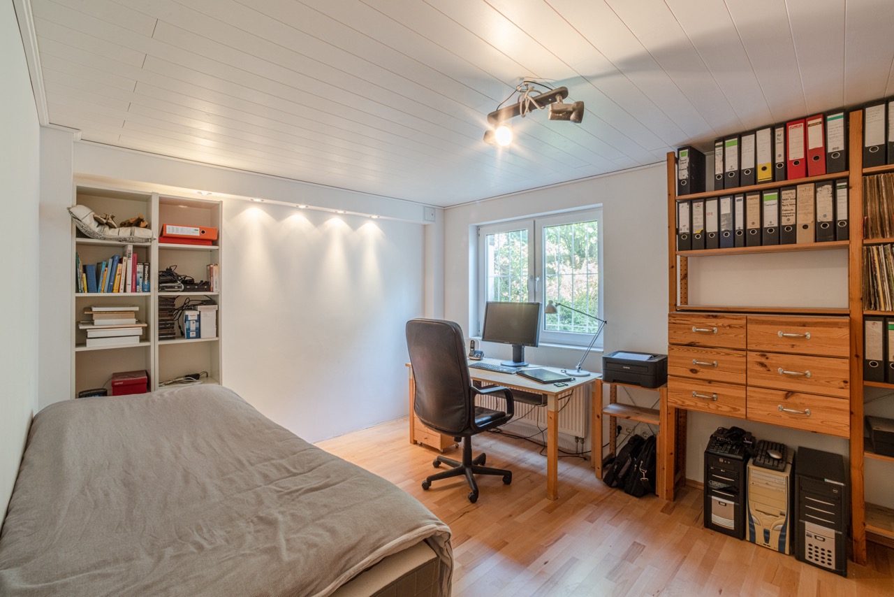 Helles Arbeitszimmer mit Schreibtisch, Regal, Bett und Ordnern, großes Fenster an der Wand.