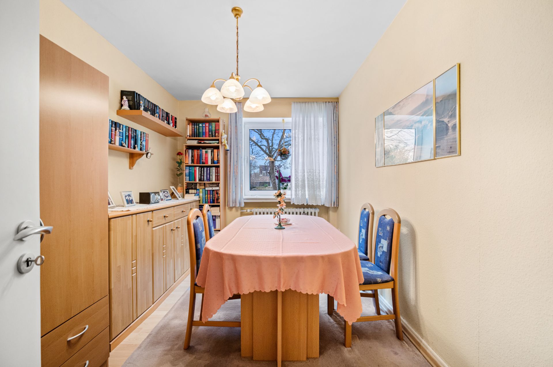 Esszimmer mit Holzmöbeln, Esstisch mit rosa Tischdecke, Bücherregalen und Fenster.