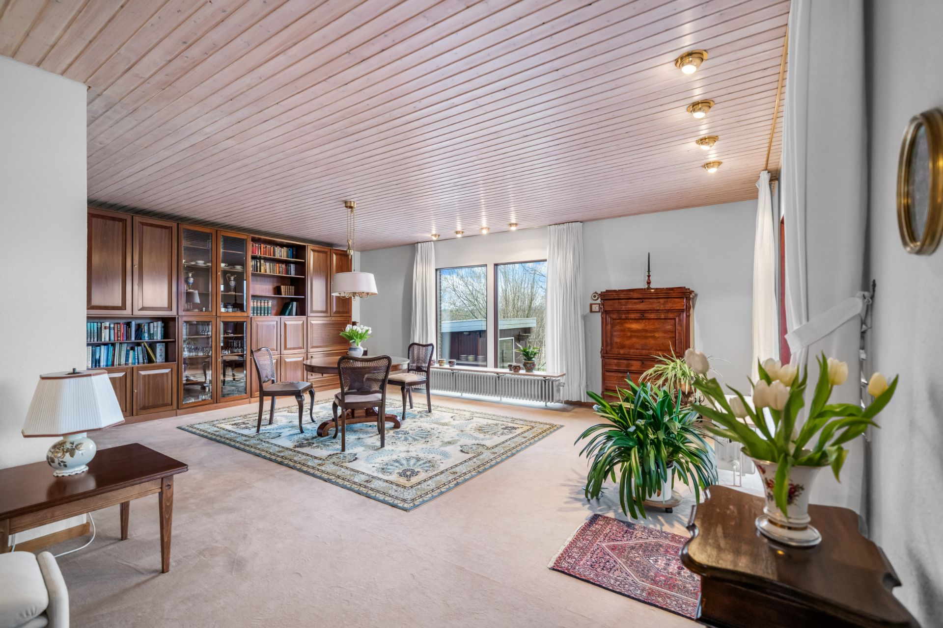 Helles Wohnzimmer mit Holzboden, Teppich, Bücherregal, Pflanzen und großen Fenstern.