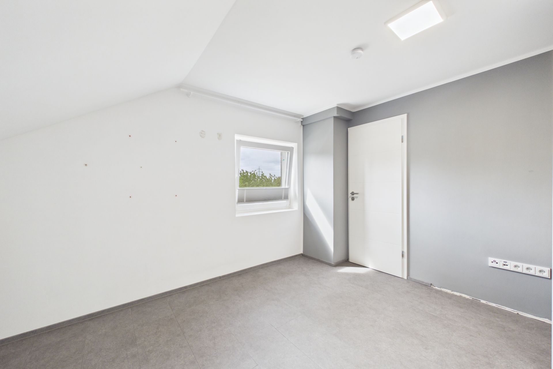 Leerer, heller Raum mit grauer Wand, Fenster und weißer Tür.