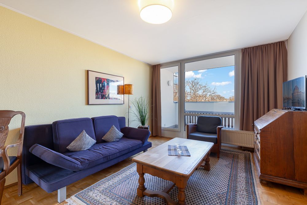 Helles Wohnzimmer mit Sofa, Sessel, Holztisch, Teppich, Schrank und Zugang zu einem Balkon.