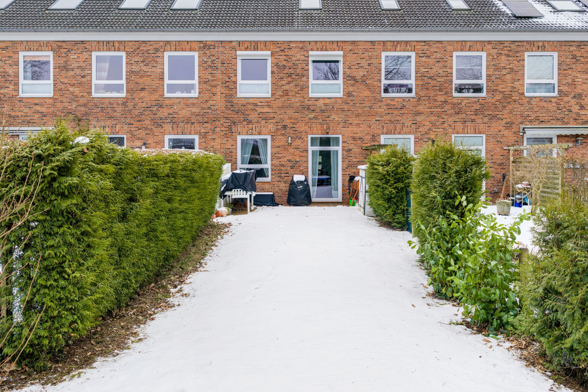 Backstein-Reihenhaus mit mehreren Fenstern, verschneiter Weg und grüne Hecken im Vorgarten.