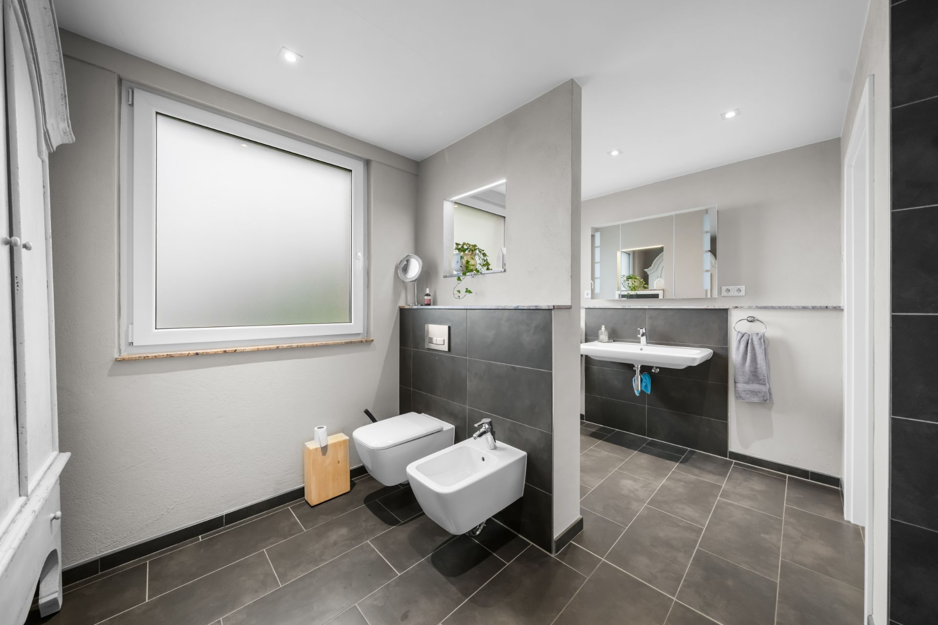 Modernes Badezimmer mit Bidet, WC, großem Fenster und Waschbecken mit Spiegel.