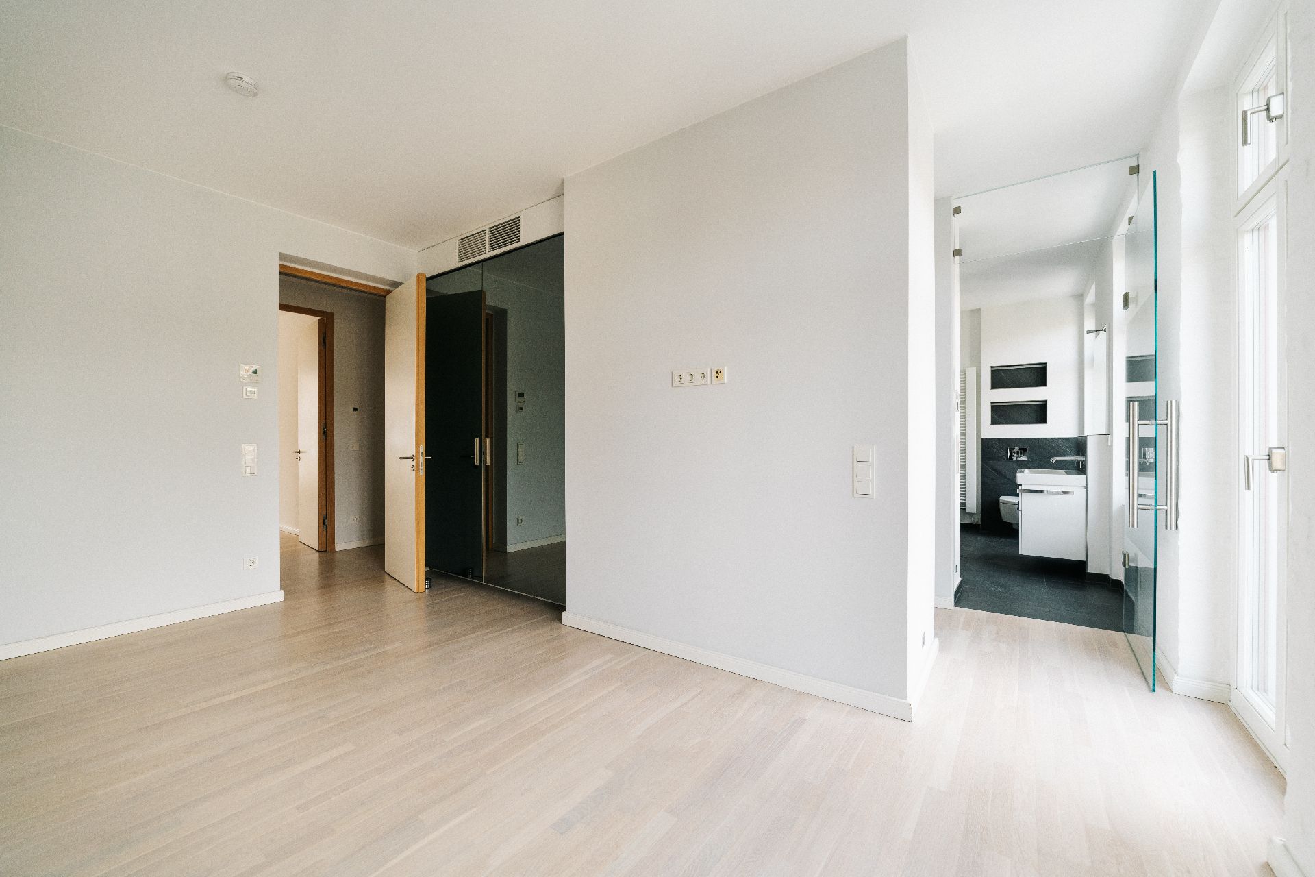 Helles, leeres Zimmer mit Holzboden und offenen Türen zu Flur und Küche.