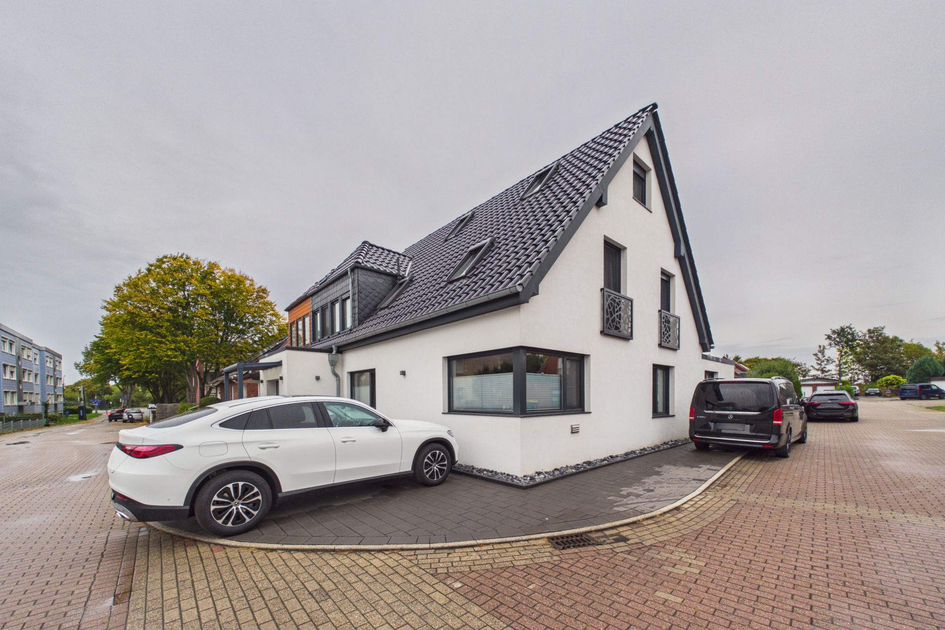 Modernes weißes Wohnhaus mit dunklem Dach, zwei Autos sind davor geparkt.