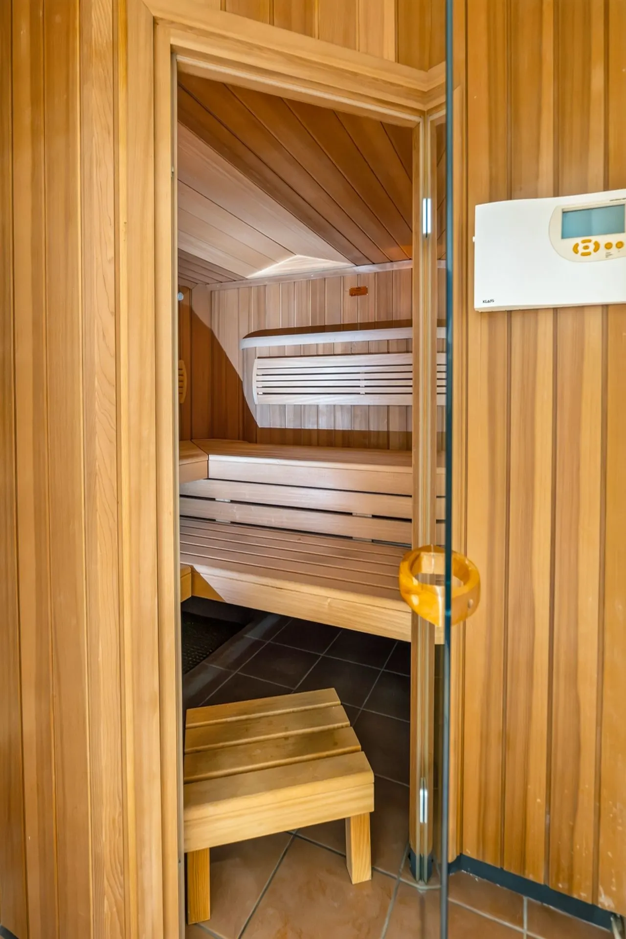 Holzsauna mit offener Tür, Holzbänken und einem kleinen Holzschemel auf Fliesenboden.