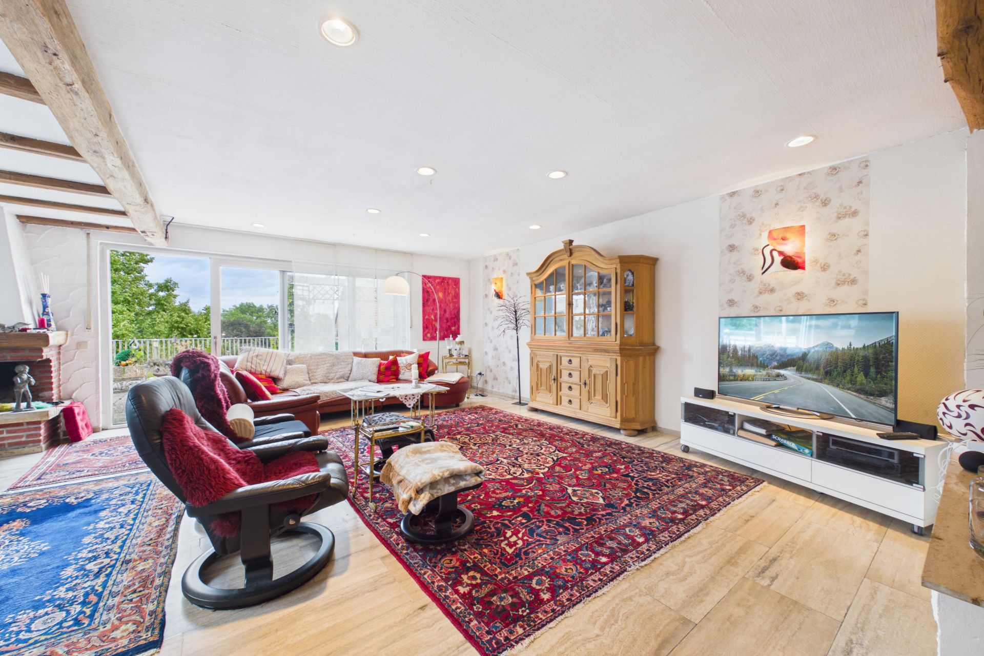 Helles Wohnzimmer mit Teppichen, Sofa, Sessel, Holzvitrine und Fernseher, große Fenster mit Blick ins Grüne.