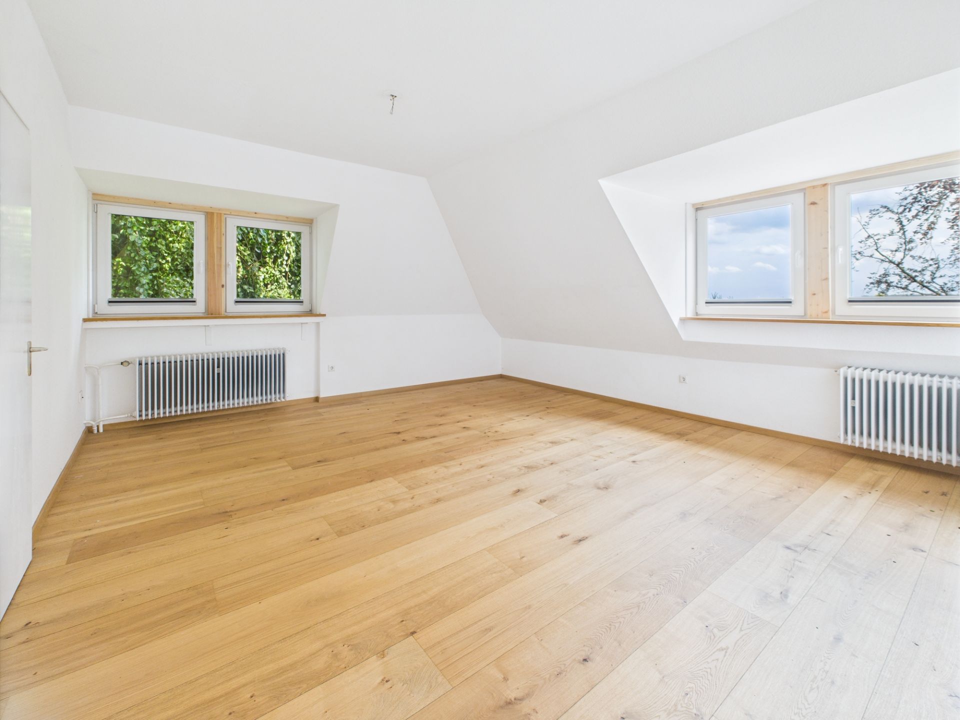 Leerer, heller Raum mit Holzfußboden und zwei Fenstern mit Heizkörpern darunter.