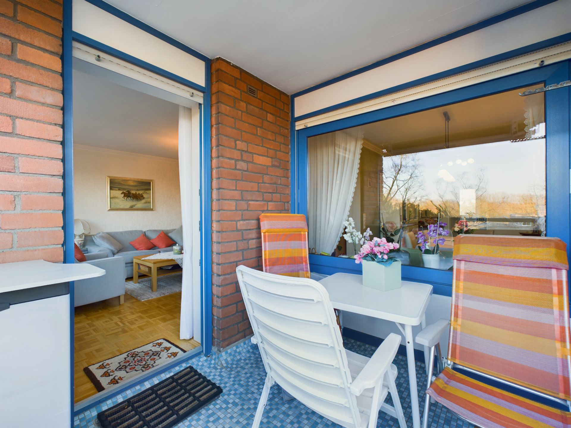 Balkon mit Tisch, zwei Stühlen und Blumen, Blick ins Wohnzimmer mit Sofa und Kissen.