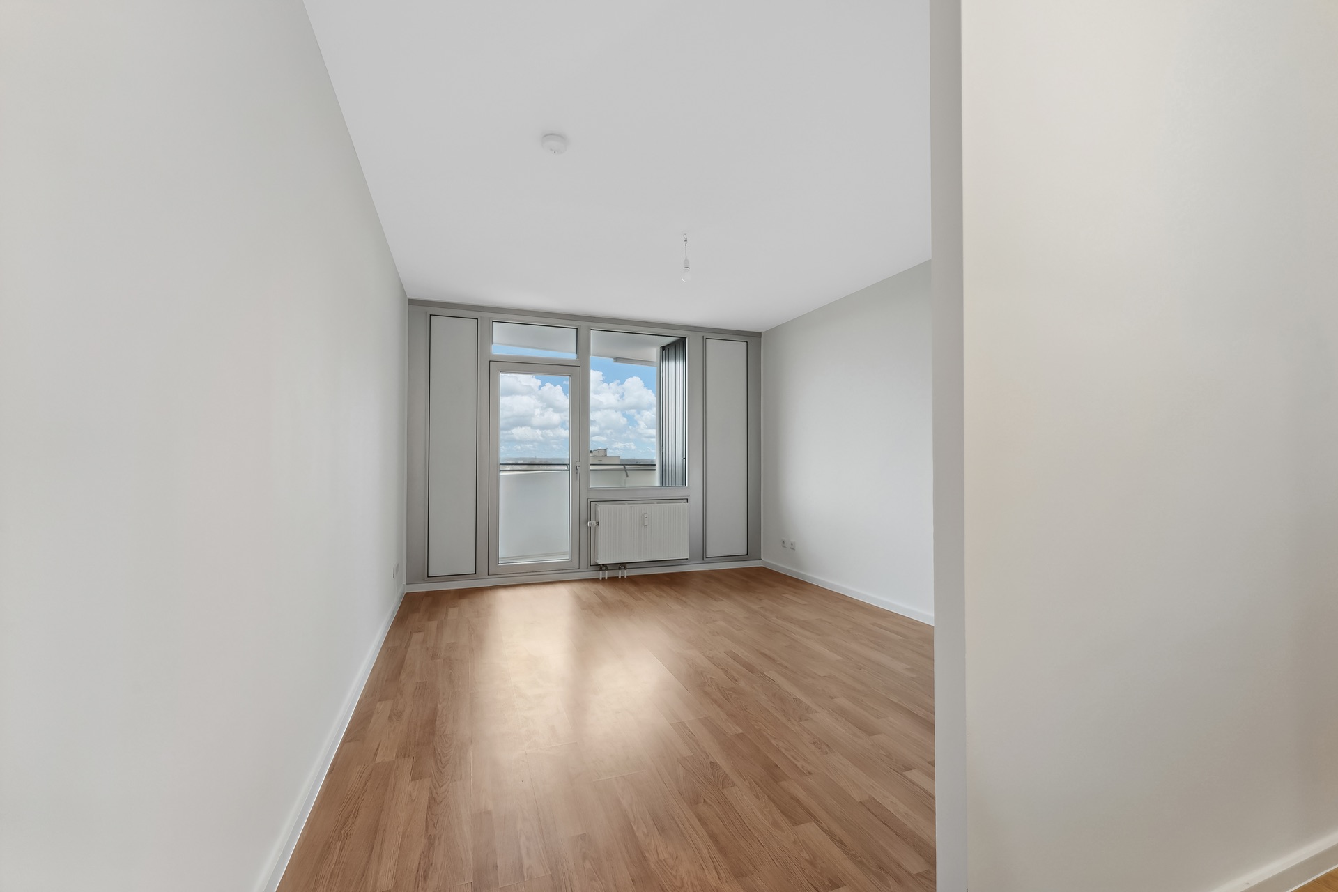 Helles, leeres Zimmer mit Holzfußboden und großem Fenster zur Terrasse.