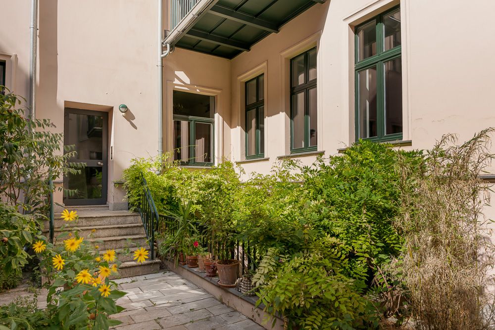 Hof mit Treppe, Grünpflanzen und gelben Blumen vor einem Gebäude mit großen Fenstern.