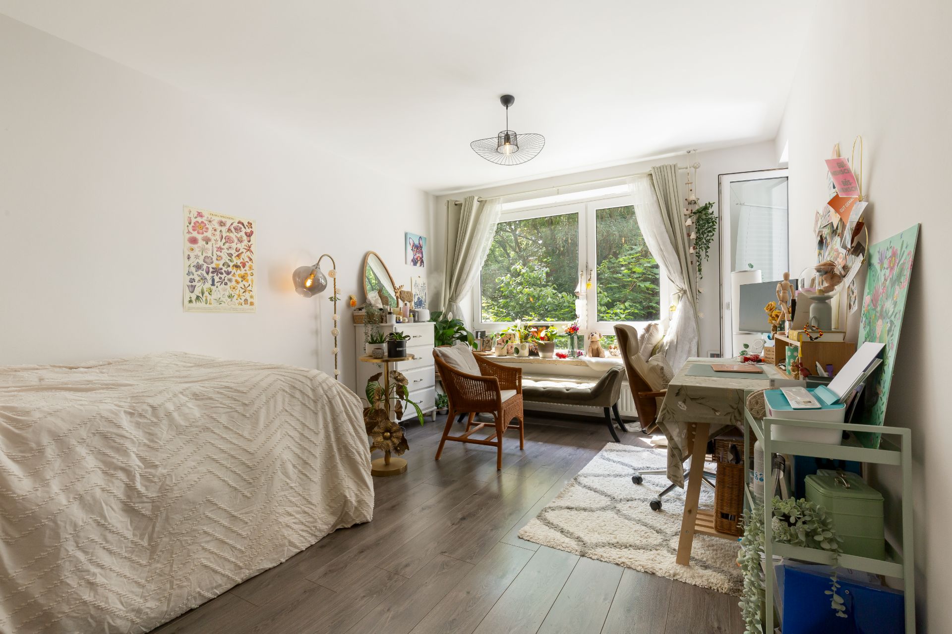 Helles Schlafzimmer mit Bett, Schreibtisch, Stuhl und großem Fenster mit Blick auf grüne Bäume.