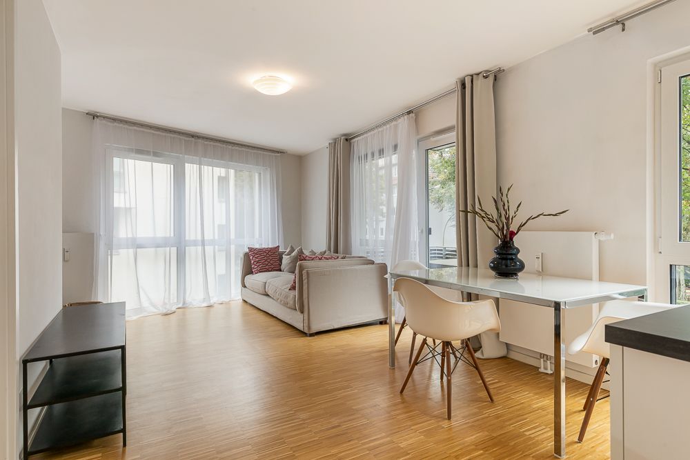 Helles Wohnzimmer mit Sofa, Esstisch, zwei Stühlen und großen Fenstern mit Vorhängen.