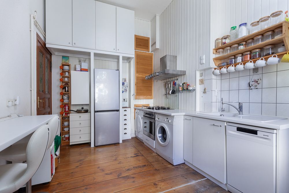 Helle Küche mit Holzboden, weißen Schränken, Geräten, offenen Regalen und einem Tisch mit Stuhl.