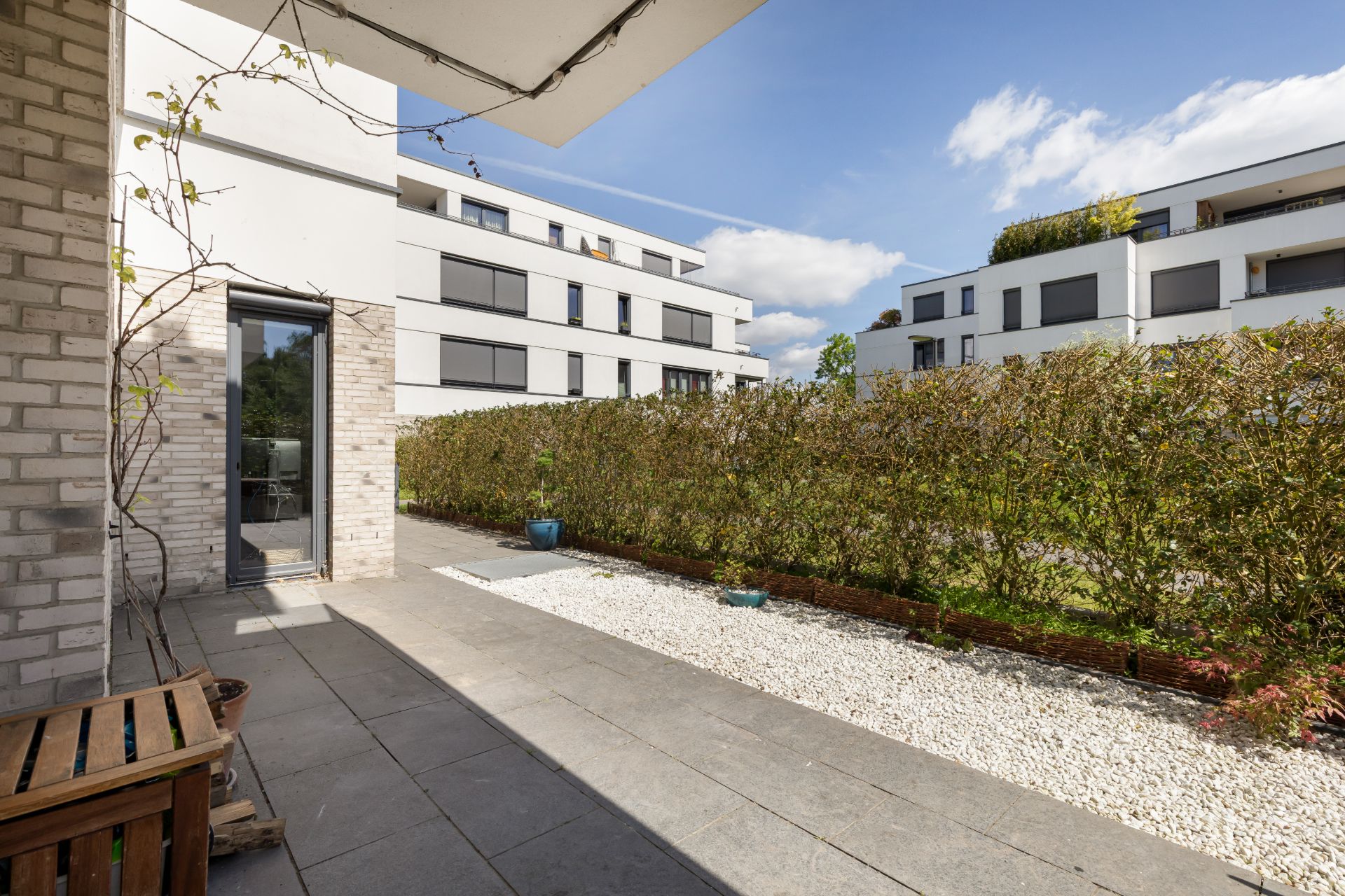 Moderne Terrasse mit Steinboden, Holzbänken, Kiesstreifen und Sichtschutzhecke.