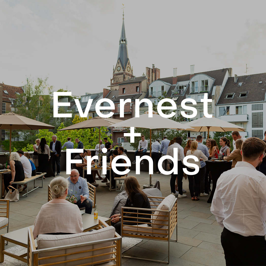 Menschen sitzen und stehen auf einer Terrasse mit Sonnenschirmen und Wohnhäusern im Hintergrund.