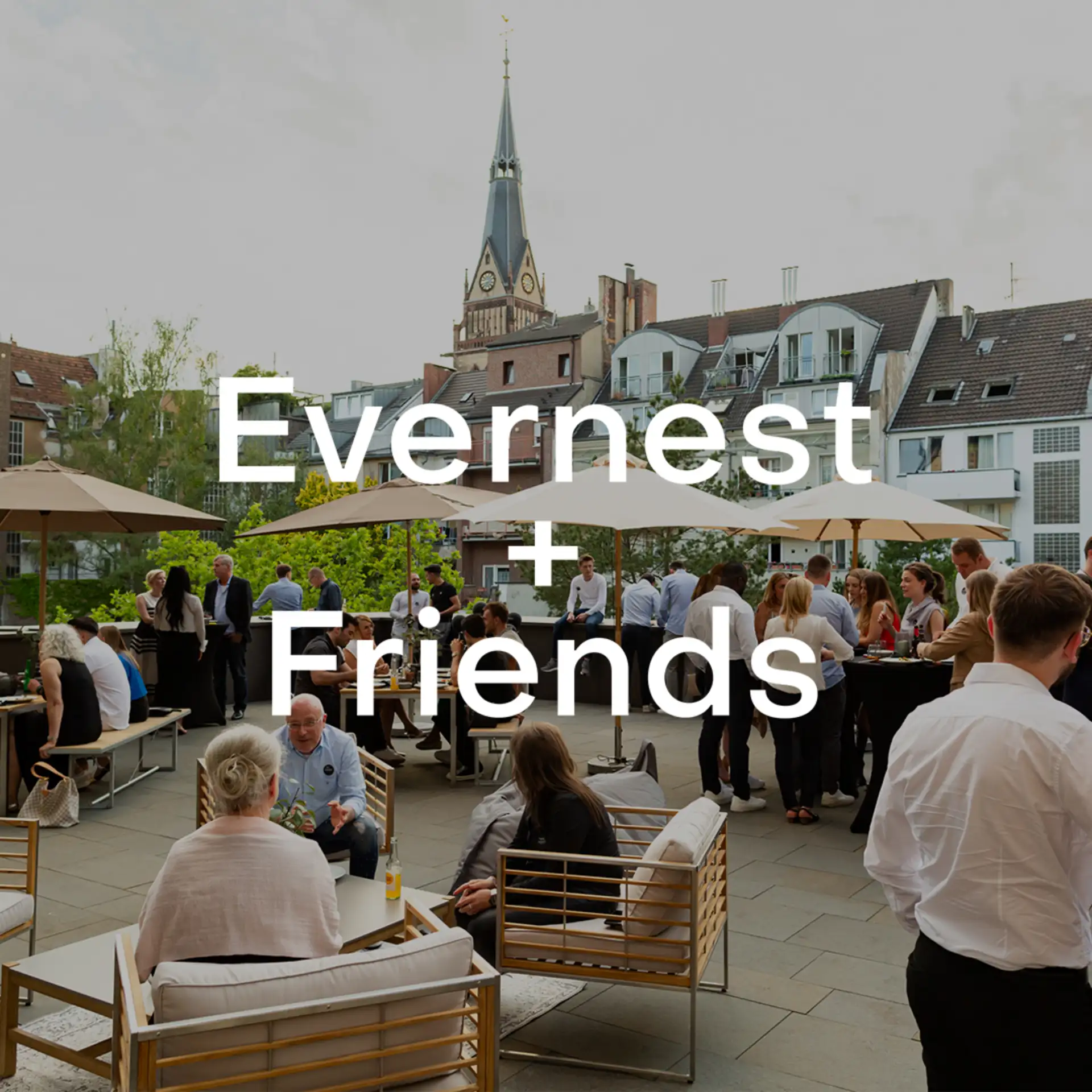 Menschen sitzen und stehen auf einer Terrasse mit Sonnenschirmen und Wohnhäusern im Hintergrund.