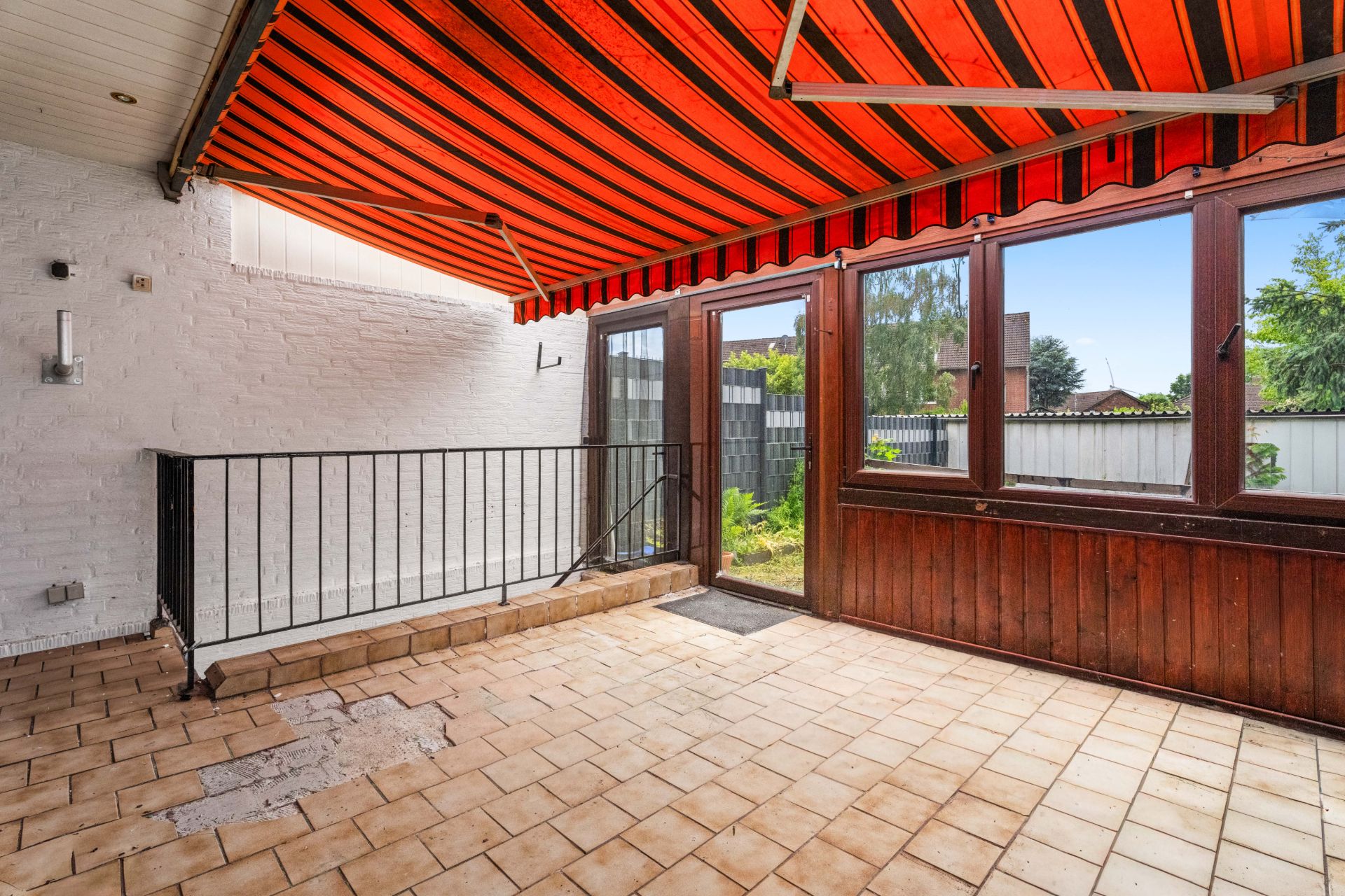 Überdachte Terrasse mit Fliesenboden, roter Markise, Geländer und Blick in den Garten.