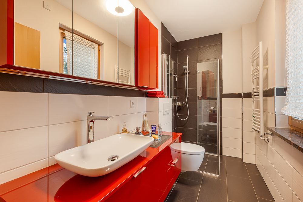 Modernes Bad mit rotem Waschbeckenunterschrank, Hänge-WC, Dusche und großem Spiegel.