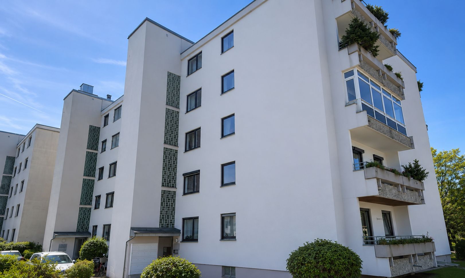 Mehrstöckiges Wohnhaus mit Balkonen und grünen Büschen davor, unter blauem Himmel.