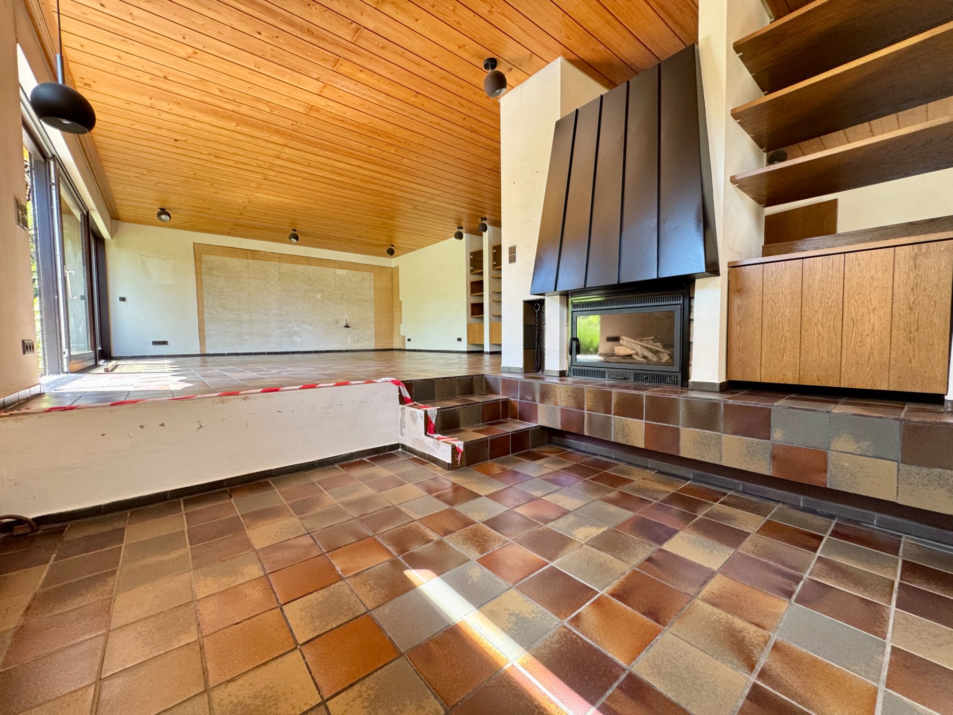 Wohnzimmer mit Kamin, gefliester Boden, Holzdecke und Regalen an der rechten Wand.