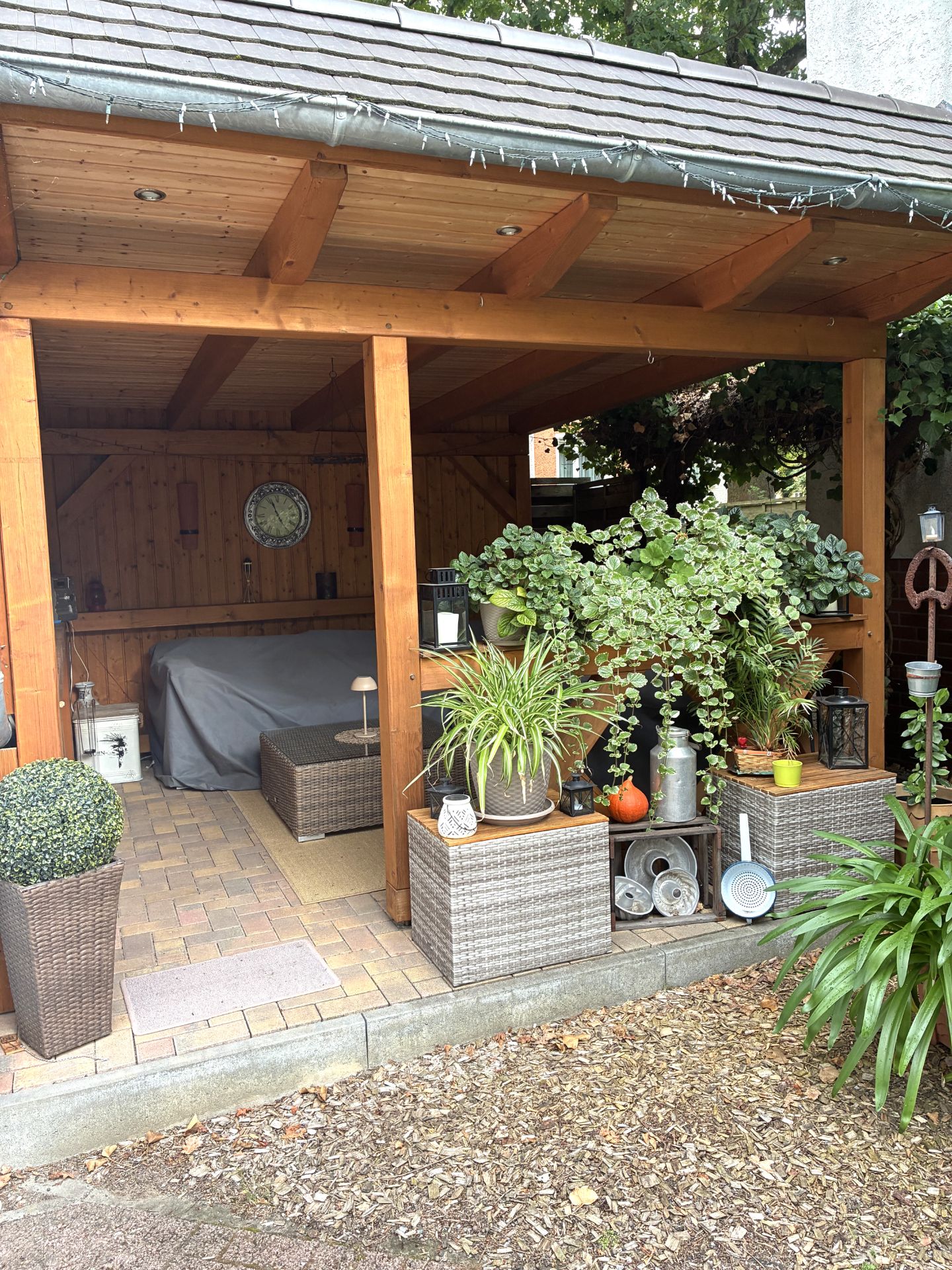 Überdachter Gartenbereich mit Holzstruktur, Pflanzen, Sitzgelegenheit und Dekorationen.