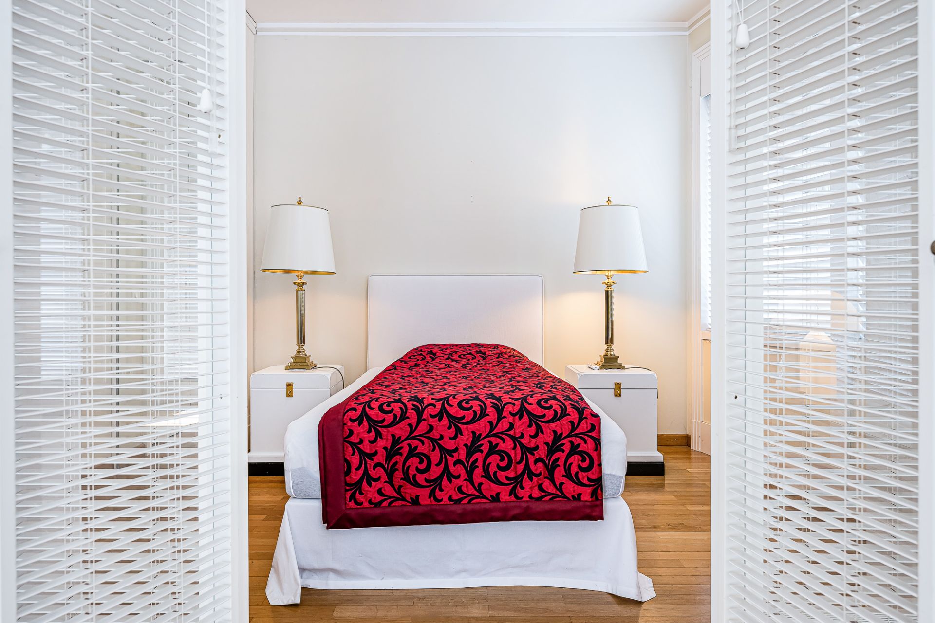 Einzelbett mit roter Decke, zwei Nachttischen und zwei Lampen in hellem Schlafzimmer.