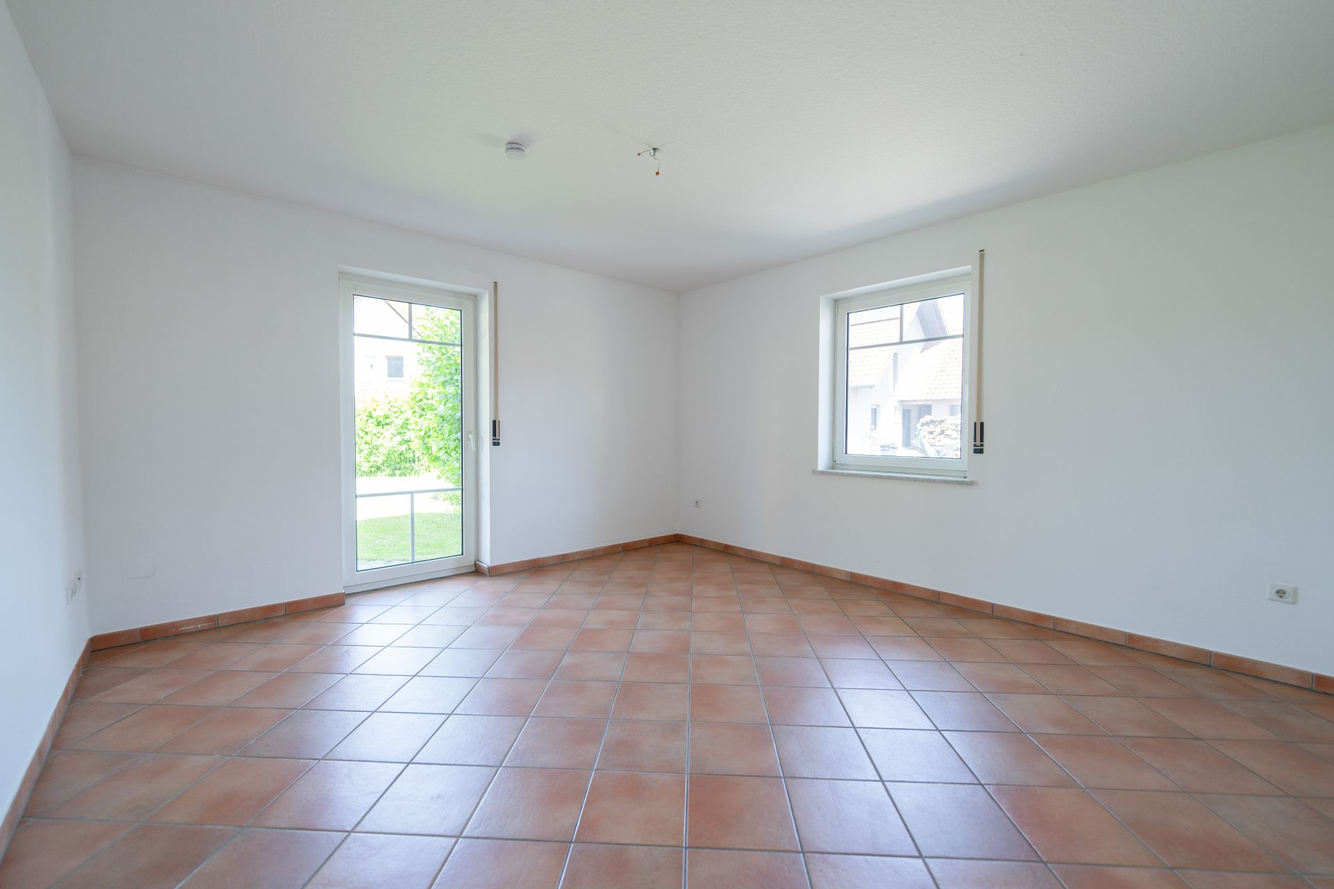 Leerer Raum mit Fliesenboden, weiß gestrichenen Wänden, Fenster und Glastür zum Garten.