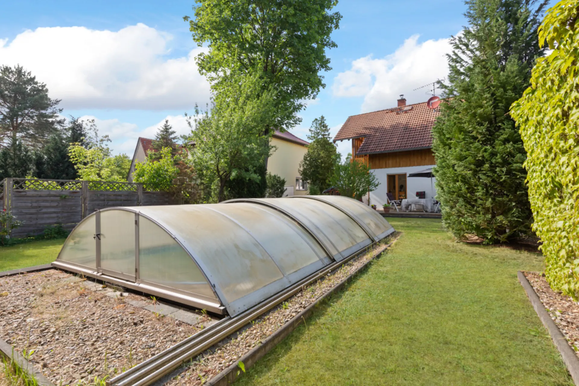 Überdachtes Schwimmbecken im Garten mit Rasen, Bäumen und Wohnhaus im Hintergrund.