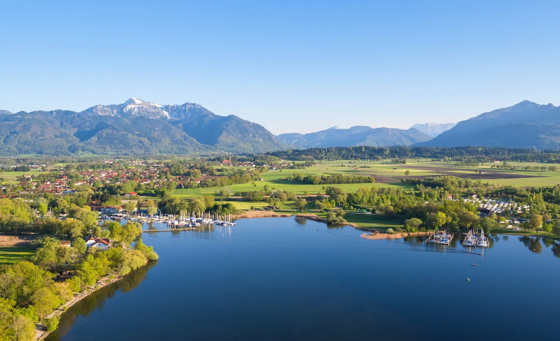 Luftaufnahme eines Sees mit Segelbooten, Wiesen und Bergen im Hintergrund.