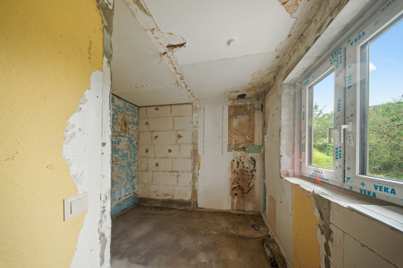 Renovierungsbedürftiger Raum mit beschädigten Wänden und offenem Fenster.