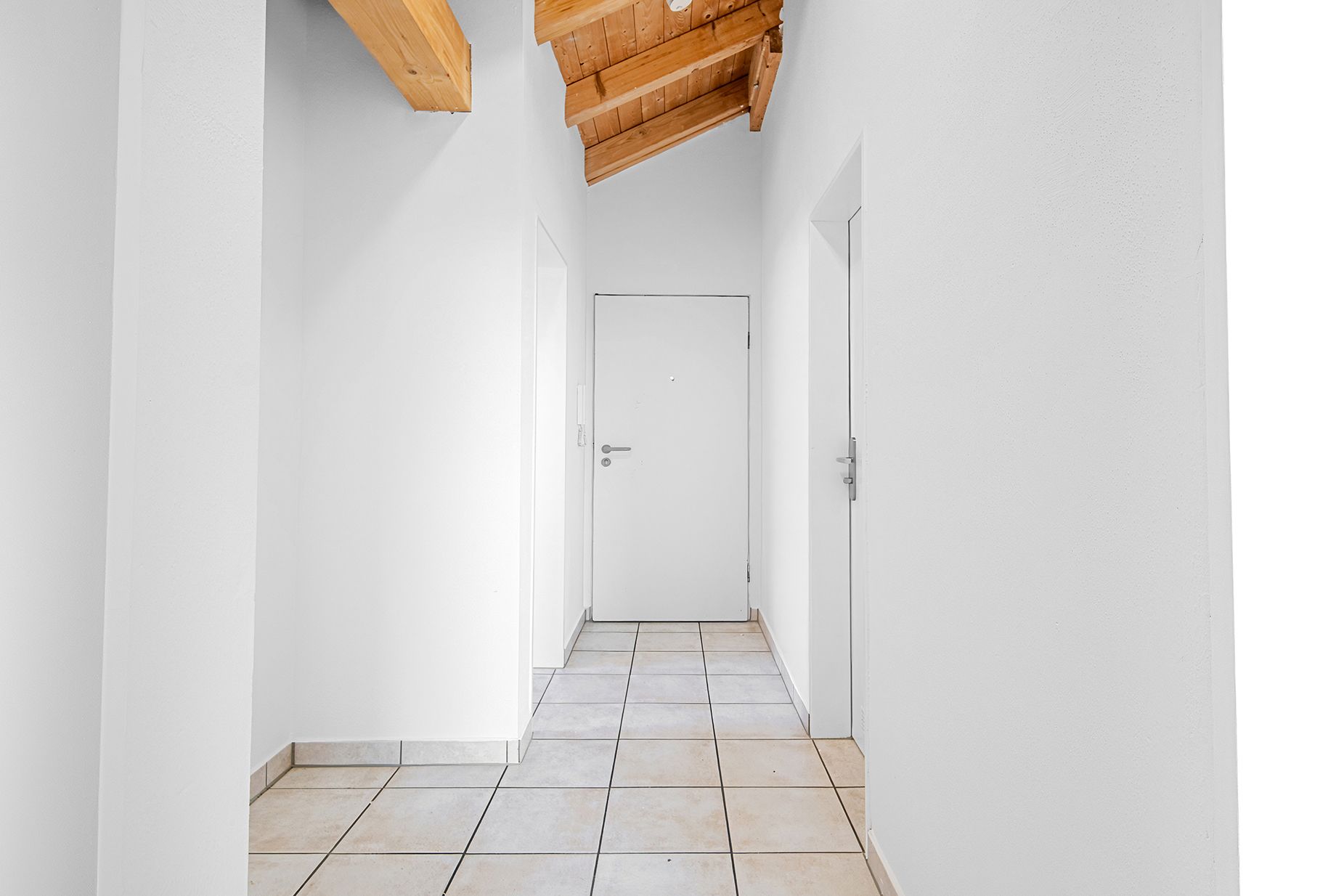 Heller Flur mit weißen Wänden, Fliesenboden und Holzbalkendecke.