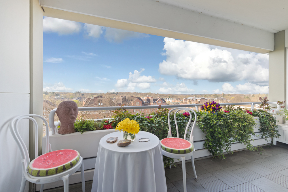 Balkon mit zwei weißen Stühlen, kleinem Tisch, Blumen und Blick auf Häuser und Himmel.