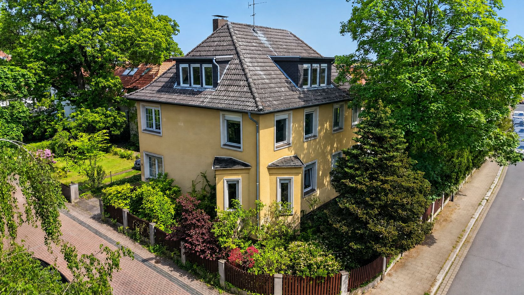 Gelbes Wohnhaus mit grauem Dach, umgeben von Garten und Bäumen an einer Straßenecke.