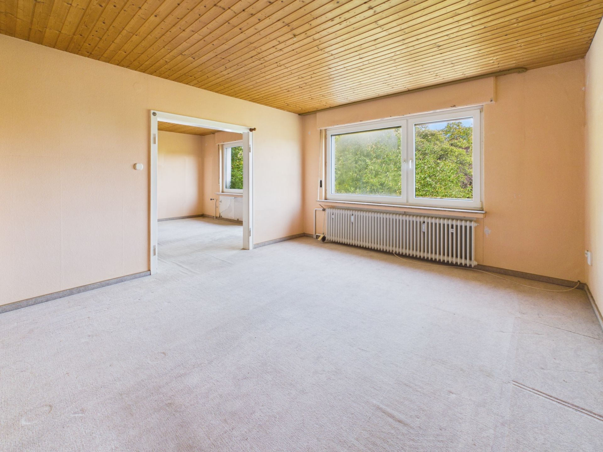 Helles, leeres Zimmer mit Holzdecke, großem Fenster und Durchgang zu einem weiteren Raum.