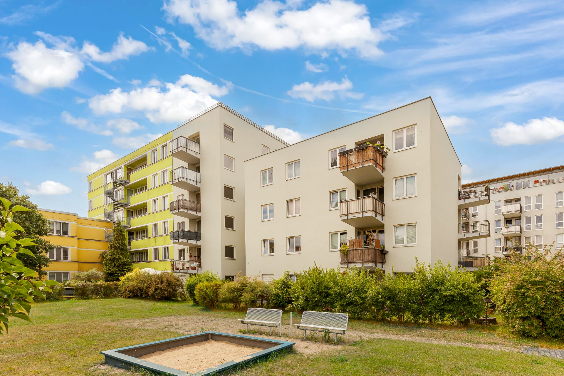 Mehrfamilienhaus mit Balkonen, davor Sandkasten und zwei Bänke auf einer Grünfläche.