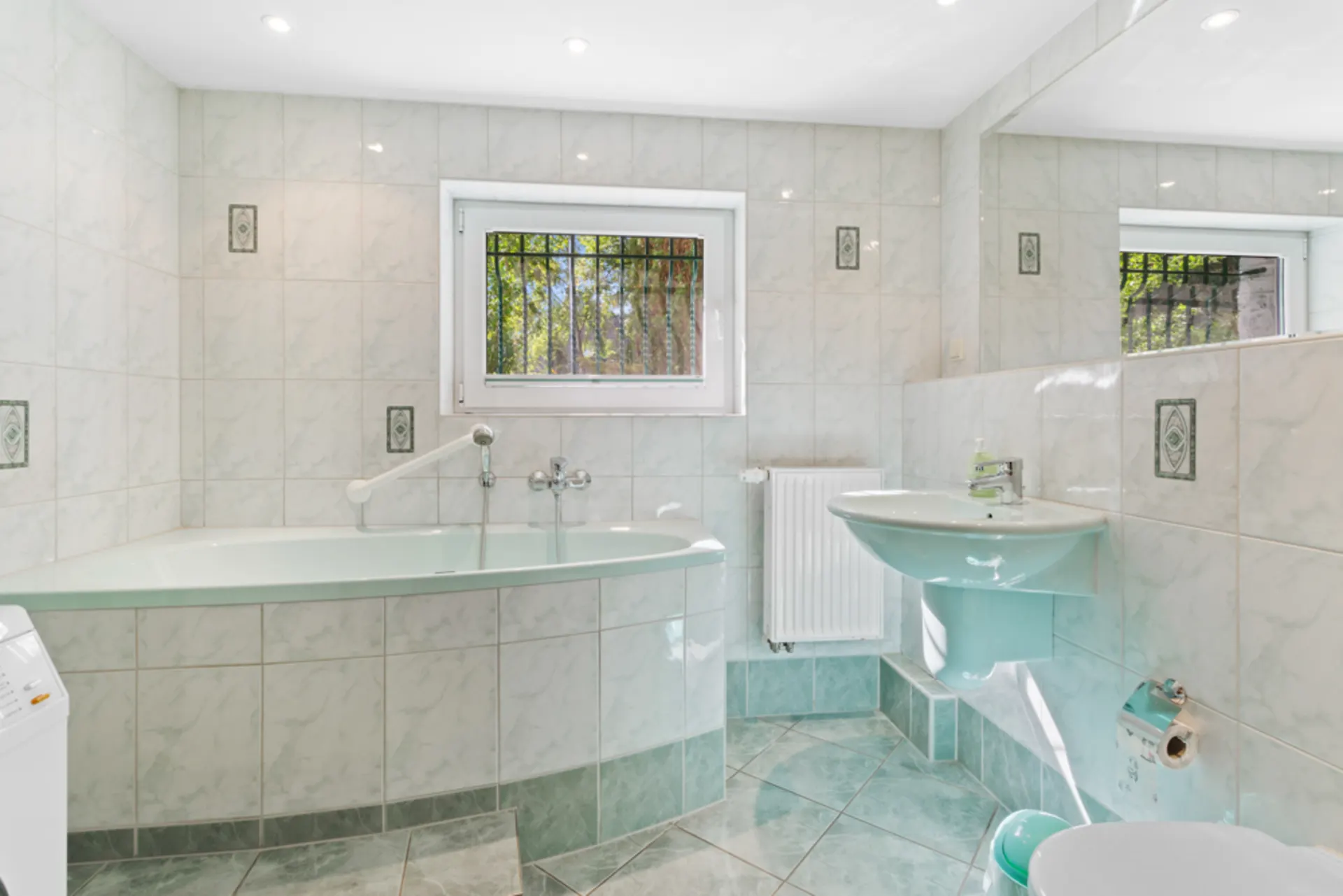 Helles Badezimmer mit Badewanne, Waschbecken, Fenster mit Gitter und gefliesten Wänden.