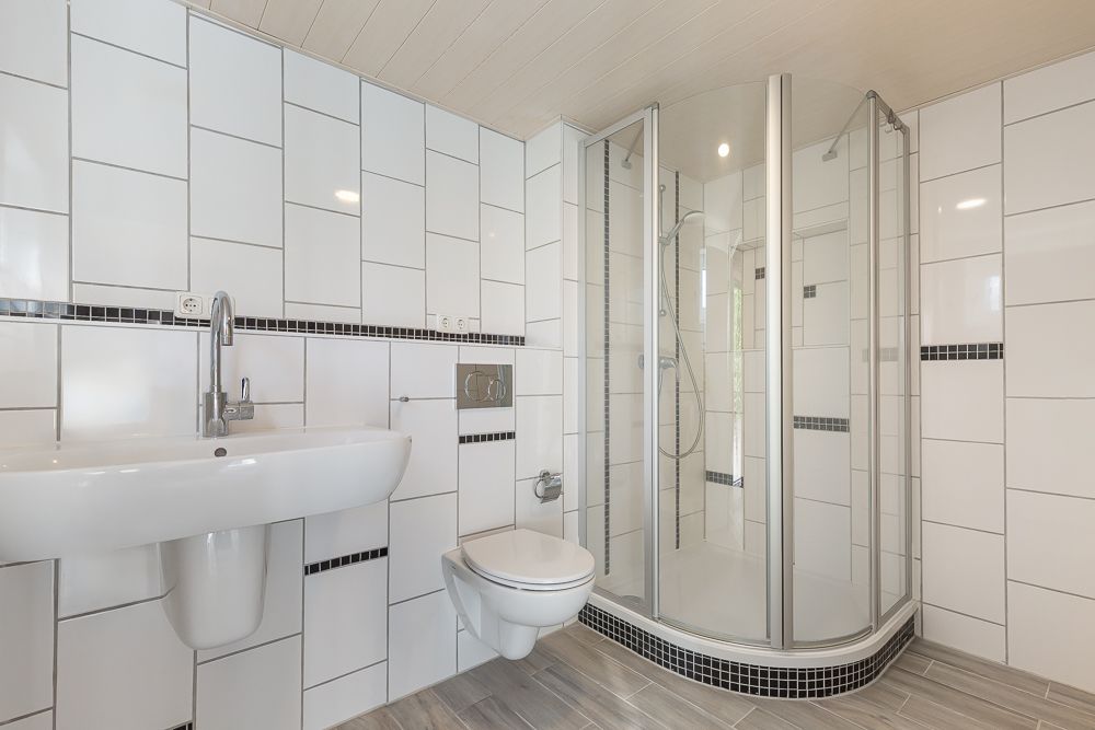 Modernes Badezimmer mit Dusche, Waschbecken und Wandtoilette, weiße Fliesen mit schwarzen Akzenten.