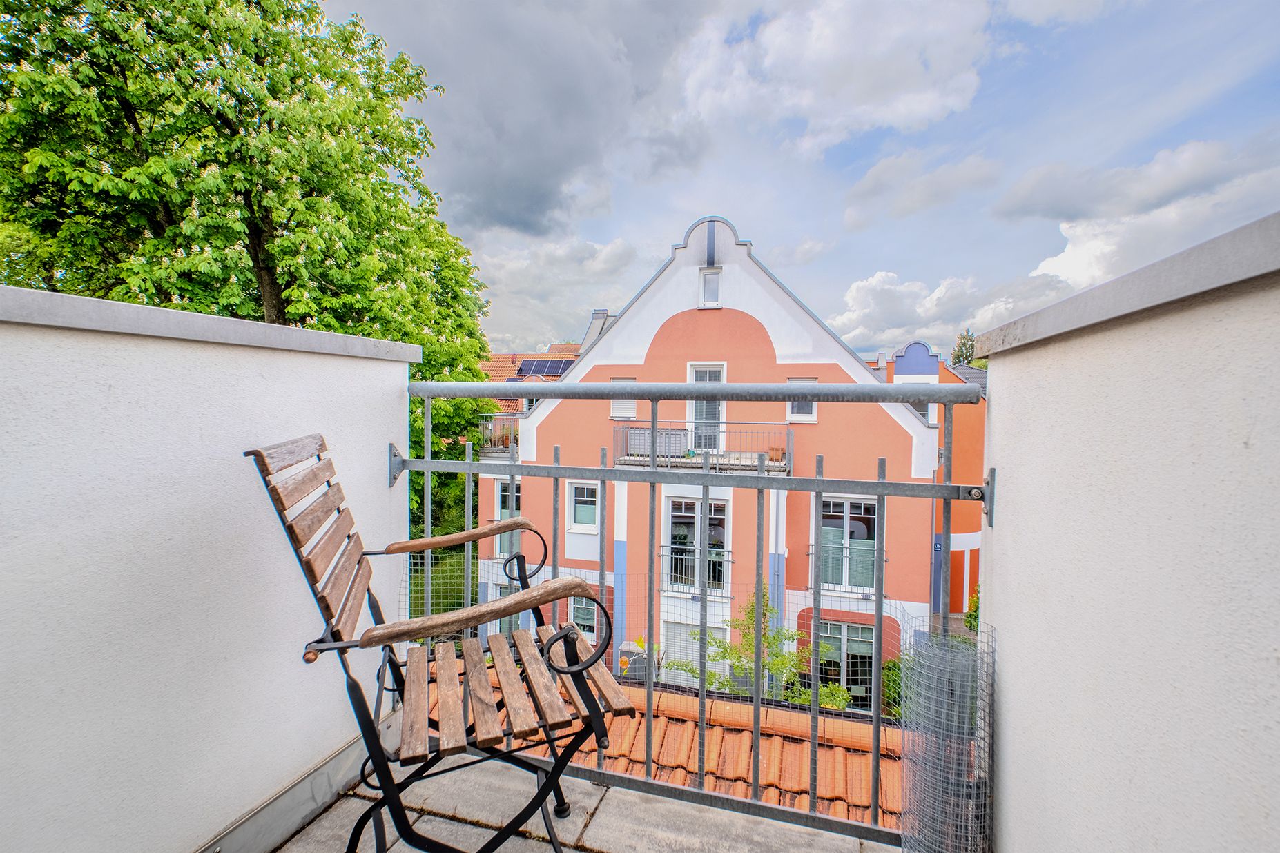 Kleiner Balkon mit einem Holzstuhl, Blick auf ein orangefarbenes Wohnhaus und Bäume.