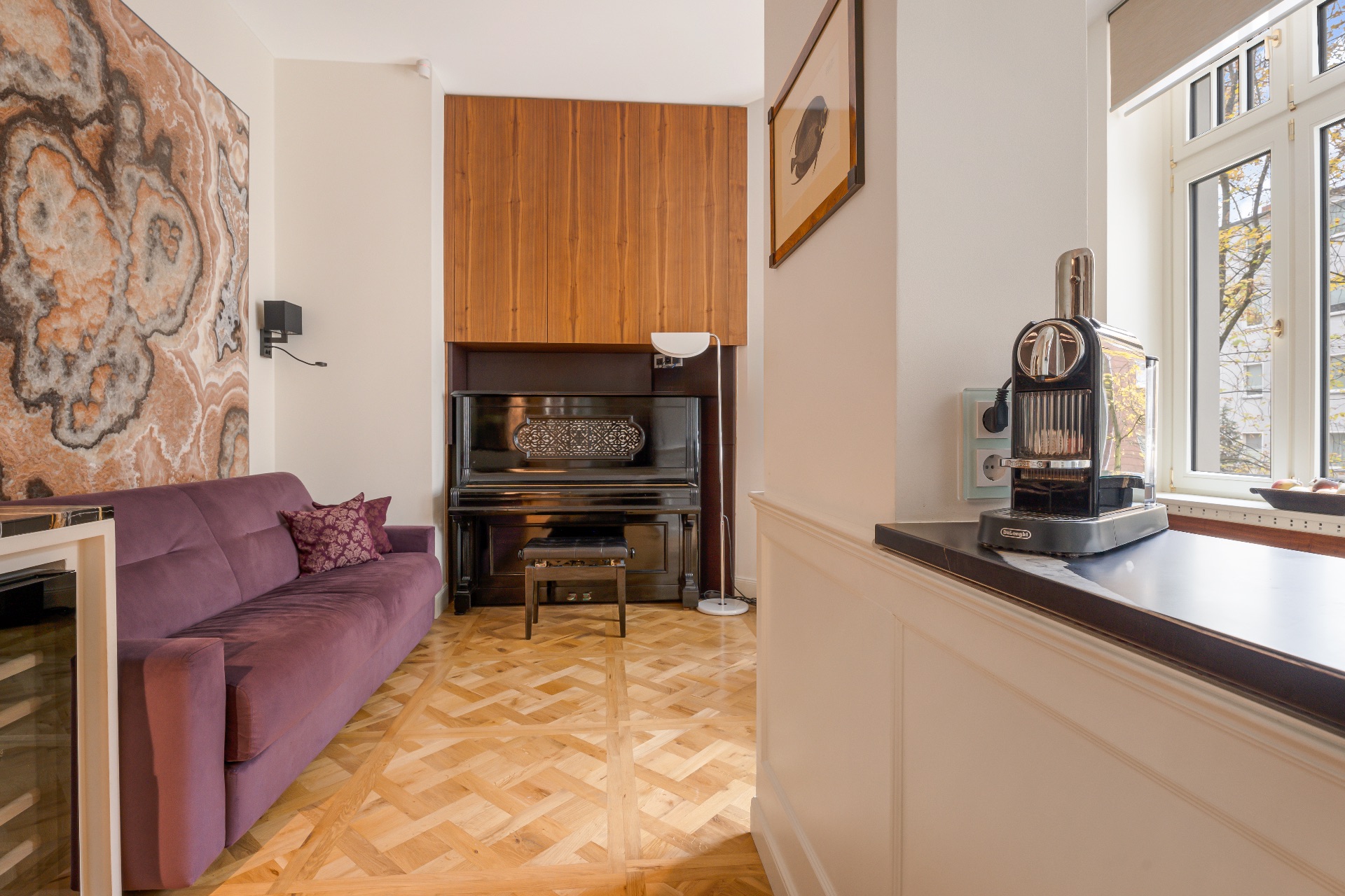 Wohnzimmer mit lila Sofa, Klavier, großem Wandbild und Fenster mit Kaffeemaschine auf der Fensterbank.
