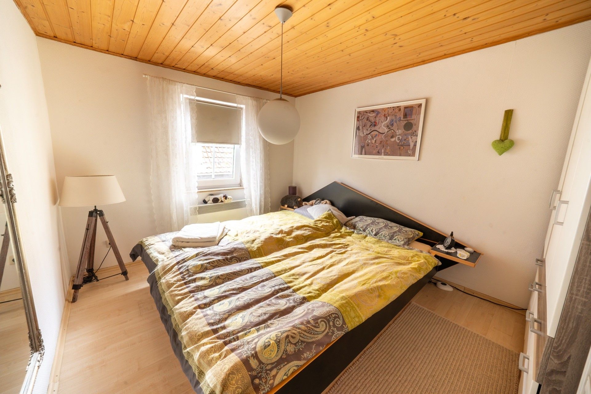 Helles Schlafzimmer mit Doppelbett, Holzdecke, Fenster, Stehlampe und Wandbild.