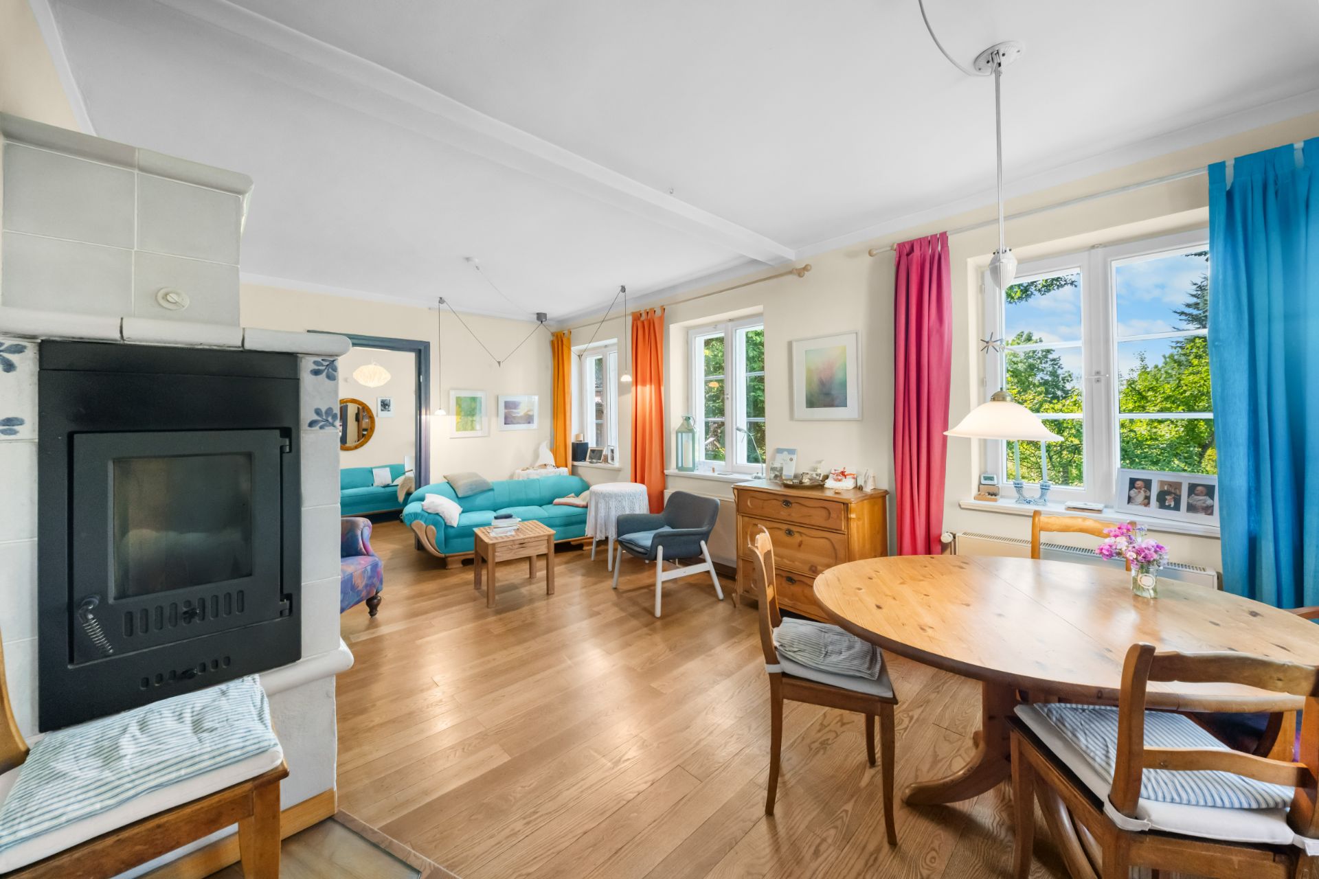 Helles Wohnzimmer mit Esstisch, bunten Vorhängen, Kamin und zwei Sofas, Holzboden und großen Fenstern.